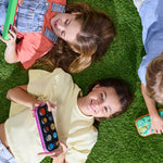 Three children lying on green artificial grass, each holding a colorful oval toy device with round buttons. The scene is shot from above, showing the children's faces, hair, and clothing in bright natural light.