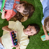 Three children lying on green artificial grass, each holding a colorful oval toy device with round buttons. The scene is shot from above, showing the children's faces, hair, and clothing in bright natural light.