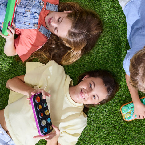 Three children lying on green artificial grass, each holding a colorful oval toy device with round buttons. The scene is shot from above, showing the children's faces, hair, and clothing in bright natural light.