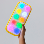 A hand holding a rectangular light-up toy with a yellow border and ten glowing multicolor buttons on a plain light gray background.