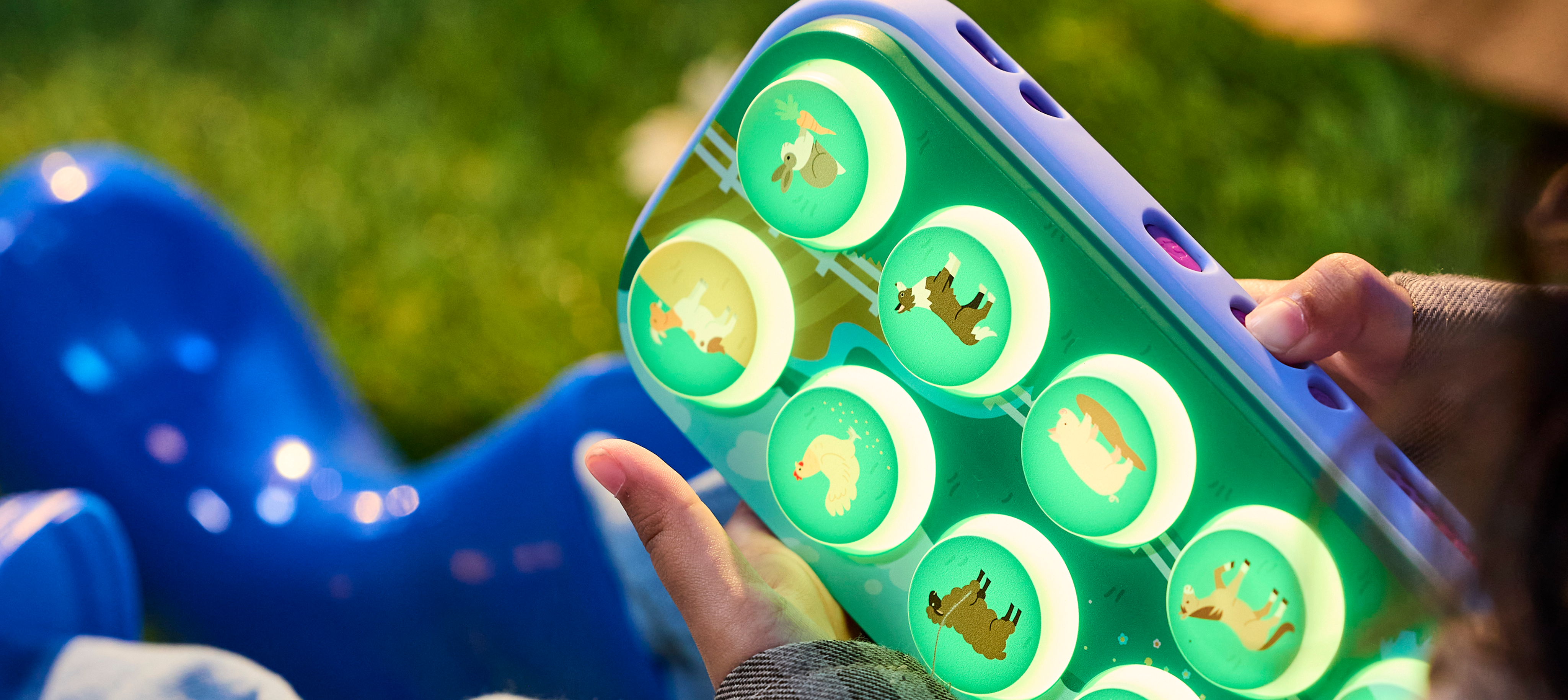 A close-up of a person holding a colorful, glowing children's toy outdoors on grass. The toy features a purple frame and multiple circular green-lit compartments with animal-shaped cutouts, including a dog, cat, and horse.