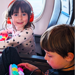 Two children on an airplane with a portable travel tablet and colorful toy.