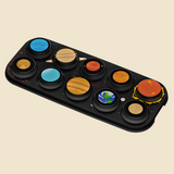 A black rectangular toy with circular sun and planet designs, including Jupiter, Saturn, Mars, Venus, Earth, and other stylized celestial motifs, shown on a plain beige background.